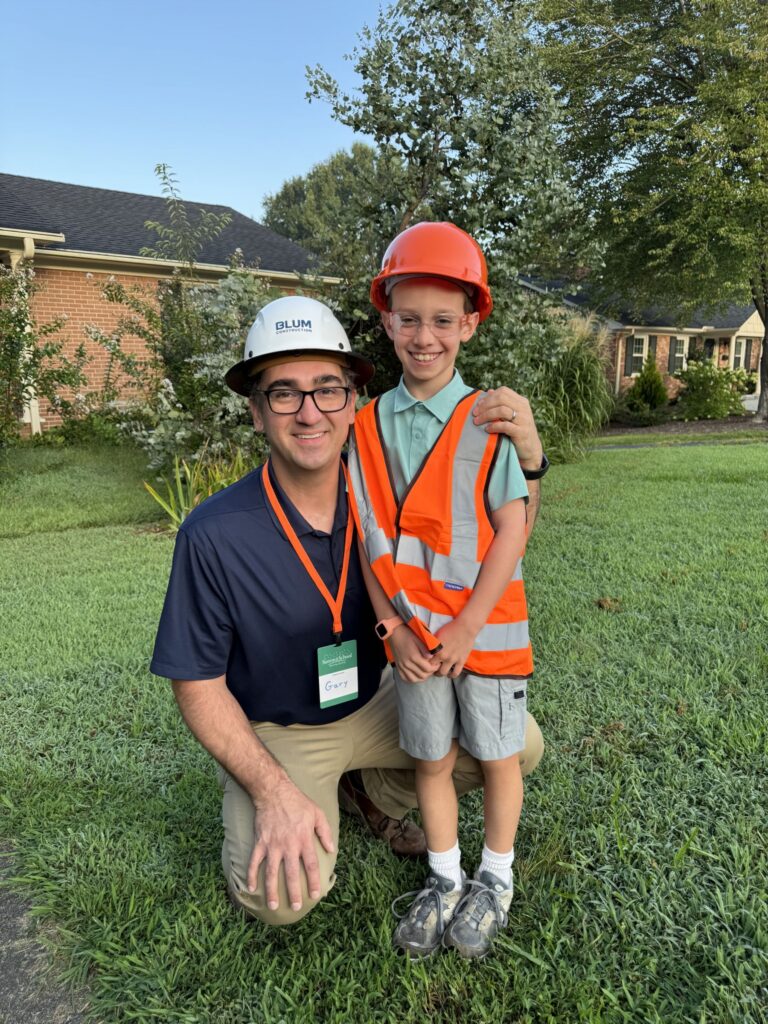Blum Construction employee wearing a company hard hat kneeling beside a smiling child in a safety vest and hard hat.