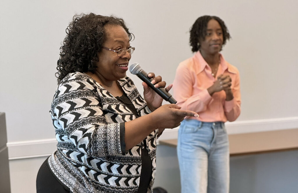 Woman speaking into a microphone during a community outreach event, with another attendee standing behind her.